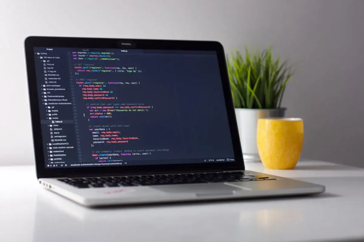 Laptop z kodem JavaScript, który pokazuje, jak otworzyć stronę internetową. Obok stoi żółty kubek i zielona roślina.